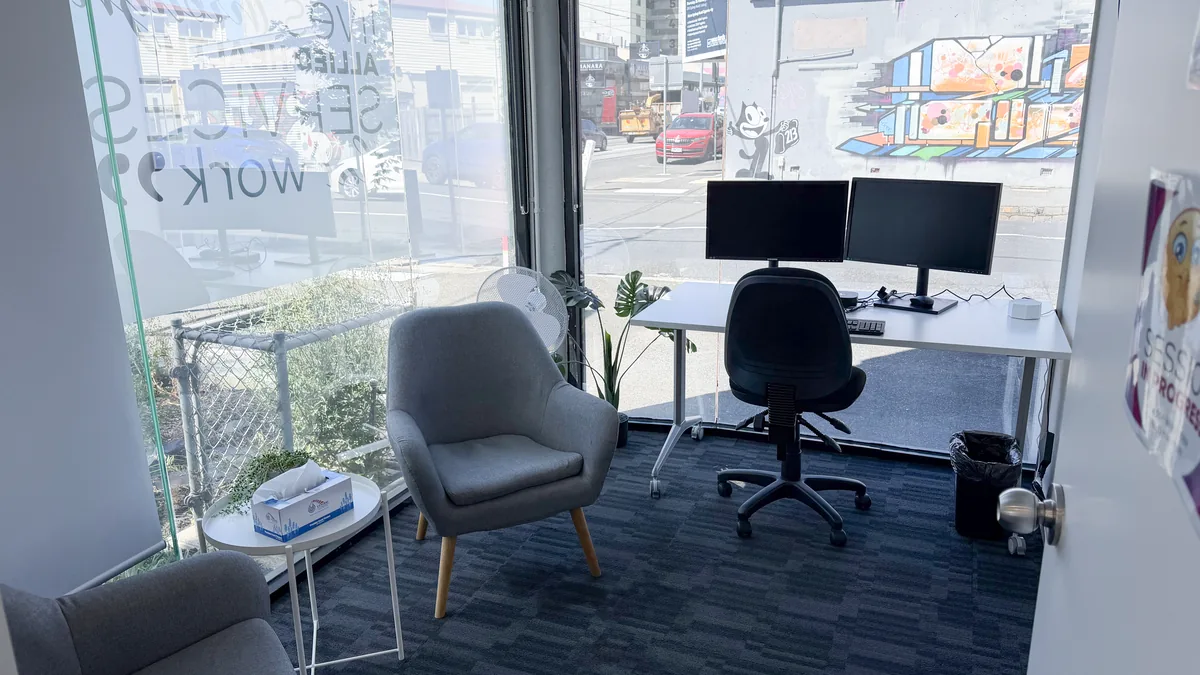 Counselling room with a soft armchair, side table and clinician desk by a bright street-facing window at ORS Coburg