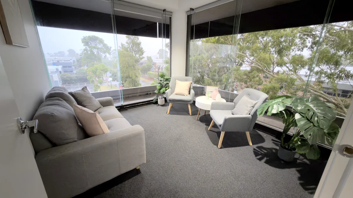 Counselling room with a lounge, two armchairs and treetop views through corner windows at ORS Mount Waverley